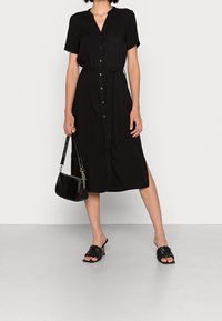 Schwarzes Hemdkleid mit kurzen Ärmeln, Knopfverschluss und Taillengürtel. Kombiniert mit einer schwarzen Texturtasche und geflochtenen schwarzen Sandalen.