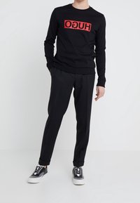 Schwarzer Kapuzenpullover mit rotem "HUGO"-Logo, kombiniert mit schwarzen, maßgeschneiderten Hosen und schwarzen Sneakern mit weißen Sohlen, die ein strukturiertes Design aufweisen.