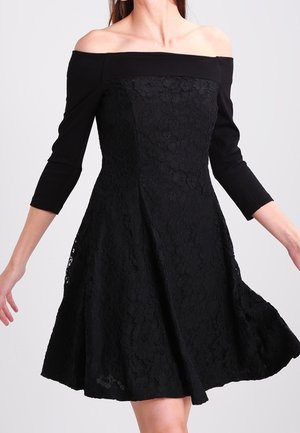 Frau trägt ein schwarzes schulterfreies Kleid mit Spitzendetails und dreiviertel Ärmeln, steht mit leicht ausgestreckten Armen.