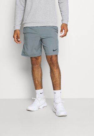 Mężczyzna ubrany w szare szorty Nike, białe skarpetki Nike oraz białe buty sportowe, stojący na tle jasnego, jednolitego tła.