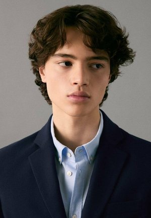 Persona joven con cabello rizado de longitud media, vistiendo una chaqueta oscura sobre una camisa azul claro con cuello, con un fondo neutro.