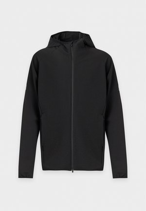 Homme portant un sweat à capuche zippé noir, un t-shirt noir, un pantalon de survêtement noir avec des bandes latérales grises, des baskets blanches et une casquette noire, debout sur fond blanc.