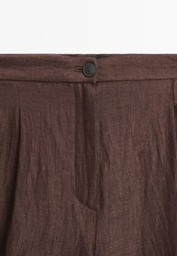 Pantalon en lin marron foncé à la taille avec un bouton noir et une fermeture éclair dissimulée, montrant la texture du tissu et les détails de la couture.