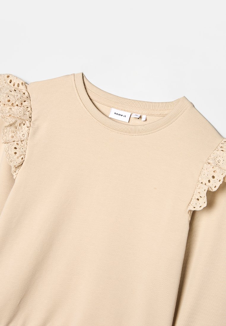 Haut beige à manches longues avec des accents de volants en dentelle sur les épaules. Fabriqué en matériau doux avec un col rond classique et une coupe décontractée.