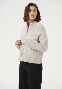 Beige zipzáras pulóver, mely texturált kötött anyagból készült, magas gallérral, bordázott mandzsettákkal és laza szabással. Fekete nadrágot viselnek alatta.