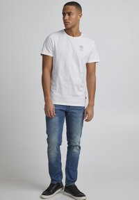 Hvit bomulls t-skjorte med rund hals og korte ermer, med et lite blått palmetre-grafikk på venstre bryst. Brukt med blå jeans og svarte joggesko.