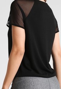 Svart T-shirt med korta, genomskinliga nätärmar. Mjukt tyg, avslappnad passform, rund halsringning och ofullbordad nederkant vid midjan.