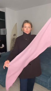 Hellrosa Schal, der vorne gehalten wird, aus weichem Stoff gefertigt. Trägt man zu einem dunkelbraunen Mantel. Im Hintergrund sind ein blaues Sofa und ein Surfbrett zu sehen.