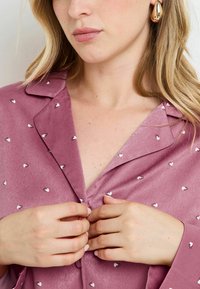 Chemise de pyjama en satin lilas avec de petits cœurs imprimés. Dotée d'un col et d'une fermeture à boutons, elle présente une texture lisse et une coupe légèrement ample.