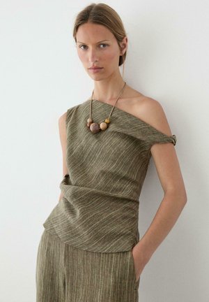 Frau trägt olivgrünes, ein-ärmeliges, strukturiertes Oberteil und passende Hose, ergänzt durch eine klobige Halskette aus Holzperlen, steht vor einer weißen Wand.