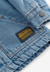 Giacca di jeans azzurra chiara con cuciture a contrasto marroni. Presenta un'etichetta nera con testo giallo, bottone in metallo e polsini ripiegati.