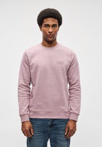 Gaiši violets sporta krekls ar garām piedurknēm un apaļu kakla izgriezumu, ar teksturētu audumu un mazu logotipa uzšuvumu uz krūtīm. Saskaņots ar zilām džinsu biksēm.