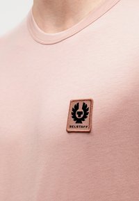 Ροζ βαμβακερό t-shirt με στρογγυλή λαιμόκοψη και υφασμάτινο καφέ μπαλώμα με μαύρο λογότυπο στην αριστερή πλευρά.