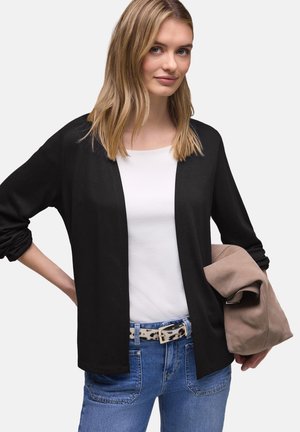 Jonge vrouw met blond haar, gekleed in een zwarte cardigan over een wit shirt en blauwe spijkerbroek, met in één hand een opgevouwen beige jas.