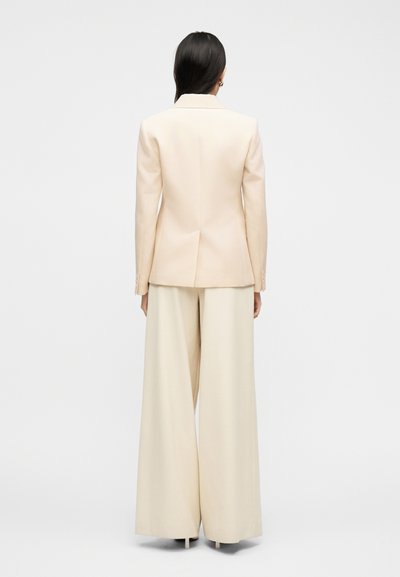 Γυναίκα με μακριά σκούρα μαλλιά, φορώντας ένα κρεμ σακάκι εφαρμοστό και παντελόνια με φαρδύ πόδι, στέκεται με την πλάτη στραμμένη προς τα εμπρός σε λευκό φόντο.