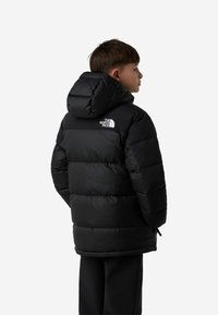 Geacă neagră matlasată, cu glugă, având un design matlasat, manșete elastice și un logo alb pe partea superioară a spatelui. Material ușor.