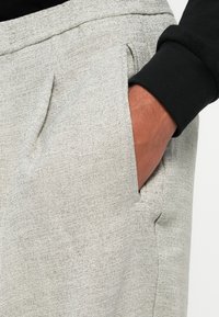 Pantalon texturé gris clair avec une poche latérale, présentant une coupe ajustée et un détail de couture visible. Haut noir à manches longues partiellement visible.