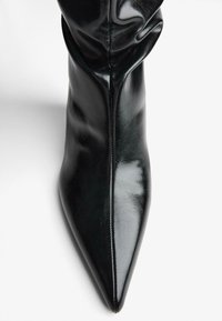 Lesklé čierne kožené čižmy so špicatou špičkou, s centrálnym švom a jemne zmršteným šachtom na bielom pozadí.