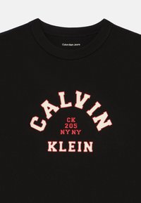 Μαύρο βαμβακερό μπλουζάκι με έντονο λευκό και κόκκινο καμπυλωτό κείμενο: "CALVIN KLEIN" και "CK 205 NY NY" κεντρικά τοποθετημένο. Κλασική στρογγυλή λαιμόκοψη.