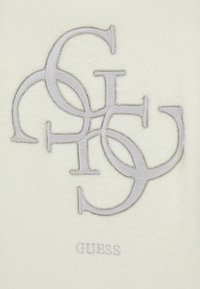 Kremo audinys su persipinančiomis iškiliosiomis pilkomis raidėmis "G" ir "S", apibrėžtomis blizgiomis metalinėmis siūlėmis, su žodžiu "GUESS" po jomis.