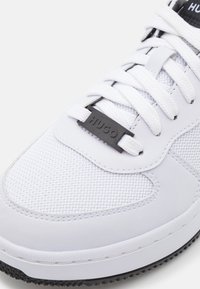 Scarpa atletica bianca con tomaia in mesh, chiusura a lacci e suola in gomma nera. Presenta un logo "HUGO" nero sul lato e accenti di design texturizzati.