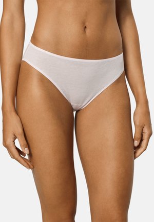 Frau trägt schlichte weiße Baumwoll-Bikini-Unterwäsche, steht mit entspannten Händen an den Seiten vor neutralem Hintergrund.