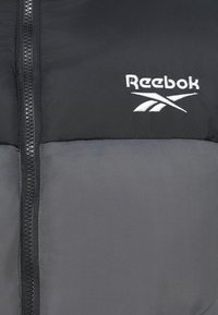 Pľacatá bunda Reebok, čierno-sivý farebný blokový dizajn, obsahuje centrálny zips a vyšívané logo v hornej časti. Hladký povrch.