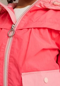 Bambino che indossa una giacca a cappuccio rosa brillante con cerniera argento e tasca bottone rosa pallido sopra una maglietta bianca.