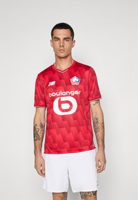 Rød atletisk trøje lavet af tekstureret stof, med et geometrisk mønster, korte ærmer, en V-hals og et hvidt logo på forsiden.