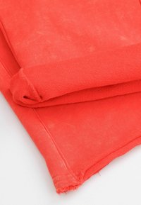 Tkanina w kolorze koralowym złożona, ukazująca teksturowaną powierzchnię. Krawędzie są lekko strzępione. Materiał wydaje się miękki i elastyczny.