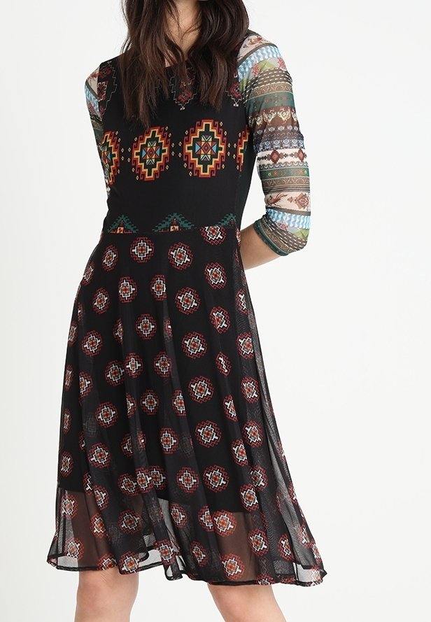 Femme portant une robe noire avec des motifs géométriques colorés et des manches longues transparentes avec des rayures multicolores.