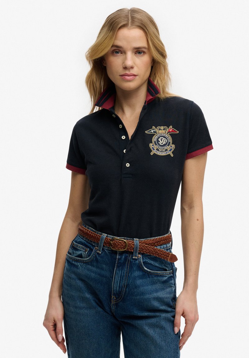 Mujer con camiseta tipo polo azul marino con cuello y emblema rojos, combinada con jeans azules y un cinturón trenzado marrón.