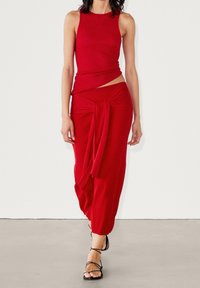 Czerwony dzianinowy zestaw składający się z dwóch części, z bezrękawną bluzką z wycięciem po boku i spódnicą z drapowaną talią związaną z przodu. Sandały na cienkich paskach w kolorze czarnym.
