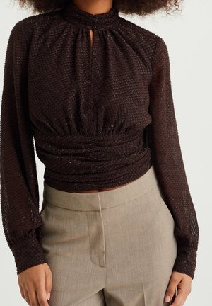 Mujer con una blusa ajustada de manga larga, texturizada, de color marrón oscuro con cuello alto y pantalones beige a medida.