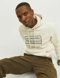 Béžová mikina s kapucí a stahovacími šňůrkami, doplněná černou nášivkou, v kombinaci s hnědými cargo kalhotami. Materiál je měkký a má volný střih.