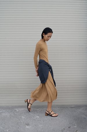 Γυναίκα που περπατάει με μακρύ ταμπά φόρεμα και μαύρα σανδάλια, με σκοτεινό πουλόβερ δεμένο γύρω από τη μέση της, μπροστά σε κλειστή γρίλια.