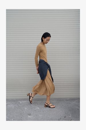 Naine kõndimas beežis pikkas kleidis ja mustades sandaalides, tal on talje ümber seotud tumedat värvi kampsun, taustal on suletud aknaroll.