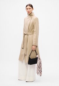 Abrigo de mezcla de lana beige con collar alto y cinturón atado. La modelo sostiene un bolso negro decorado con una bufanda de patrón rosa. Pantalones anchos blancos.