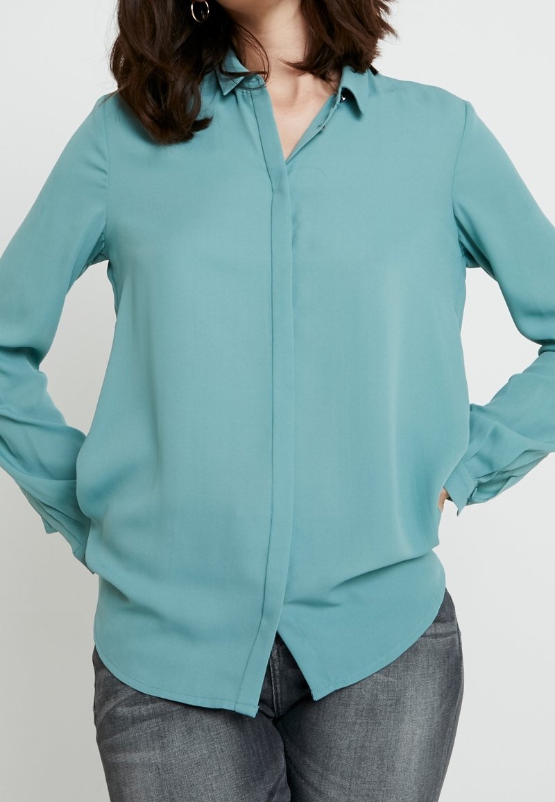 Femme portant un chemisier à manches longues bleu sarcelle avec des boutons cachés, associé à un jean noir délavé, les mains posées sur les hanches, sur fond blanc.