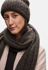 Ciemnobrązowa, teksturowana czapka beanie i szalik z gładkim wykończeniem, w zestawie z jasnobeżową kurtką i ciemną bluzką. Materiał zwraca uwagę swoją miękkością.