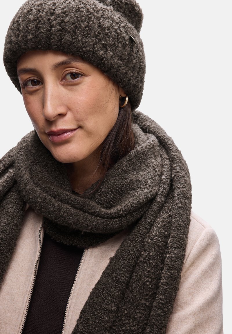 Ciemnobrązowa, teksturowana czapka beanie i szalik z gładkim wykończeniem, w zestawie z jasnobeżową kurtką i ciemną bluzką. Materiał zwraca uwagę swoją miękkością.