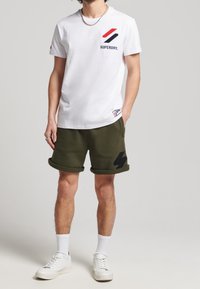 T-shirt bianca in cotone con logo e accenti rossi/blue, abbinata a pantaloni corti in pile verde oliva con motivo a saetta nera. Sneakers bianche.