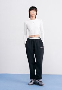 Top corto blanco de manga larga con un pequeño logo negro de Nike, combinado con pantalones de chándal negros que tienen una cintura de patrón de colores y logo.