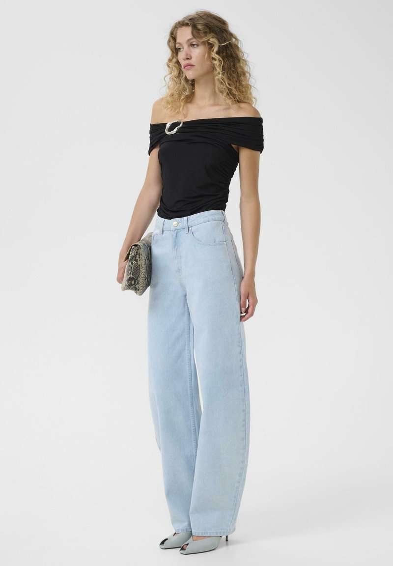 Vrouw met krullend haar die een zwarte top met blote schouders en gesp draagt, lichtblauwe wijd uitlopende spijkerbroek, lichtblauwe hakken en een clutch van slangenleer vasthoudt.