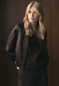 Bruine pilotjas met een gladde textuur en ritsluiting, gelaagd over een donker gebreide trui, gecombineerd met bijpassende donkere broek.