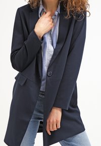 Marineblauer langer Blazer aus weichem Material mit einem einzigen Knopfverschluss, Reverskragen, seitlichen Taschen und einer lässigen Passform, getragen über einem gestreiften Hemd.
