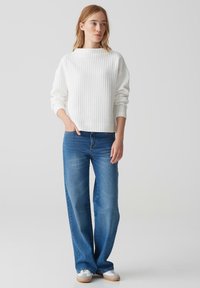 Weißer gerippter Pullover mit einem Bootsausschnitt, kombiniert mit blauen Weitbein-Jeans und weißen Sneakers mit beigen Akzenten, vor einem hellen Hintergrund präsentiert.