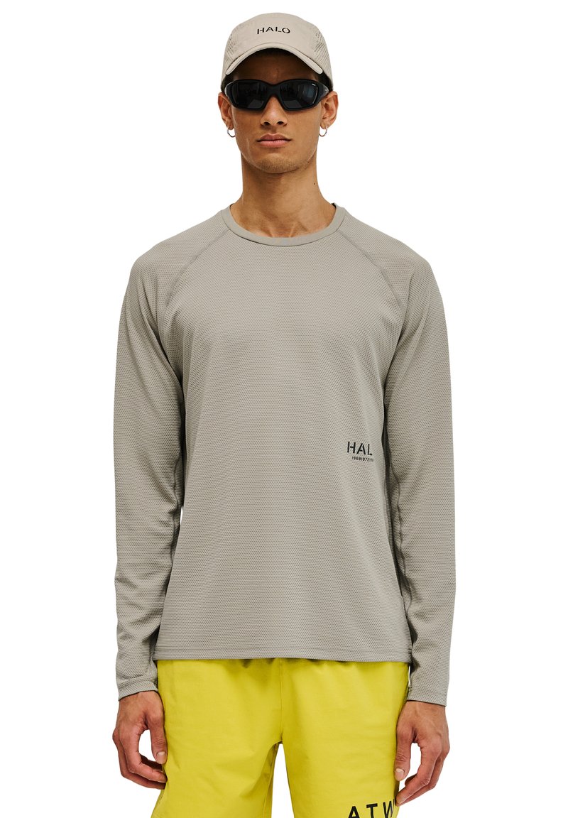 Mężczyzna w jasnoszarej koszuli z długim rękawem, jasnoszarej czapce z napisem „HALO”, czarnych okularach przeciwsłonecznych, żółtych szortach z czarnym napisem, stojący na białym tle.