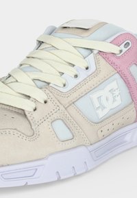 Μπεζ και ροζ αθλητικό παπούτσι DC με λευκά κορδόνια, πλέγματα και λευκή καουτσούκ σόλα, που δείχνει πλευρικές και πάνω λεπτομέρειες σε λευκό φόντο.