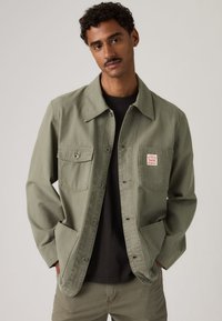 Muž s knírkem v olivově zelené bundě na zapínání s čtyřmi kapsami a černém tričku stojí před jednobarevným pozadím.